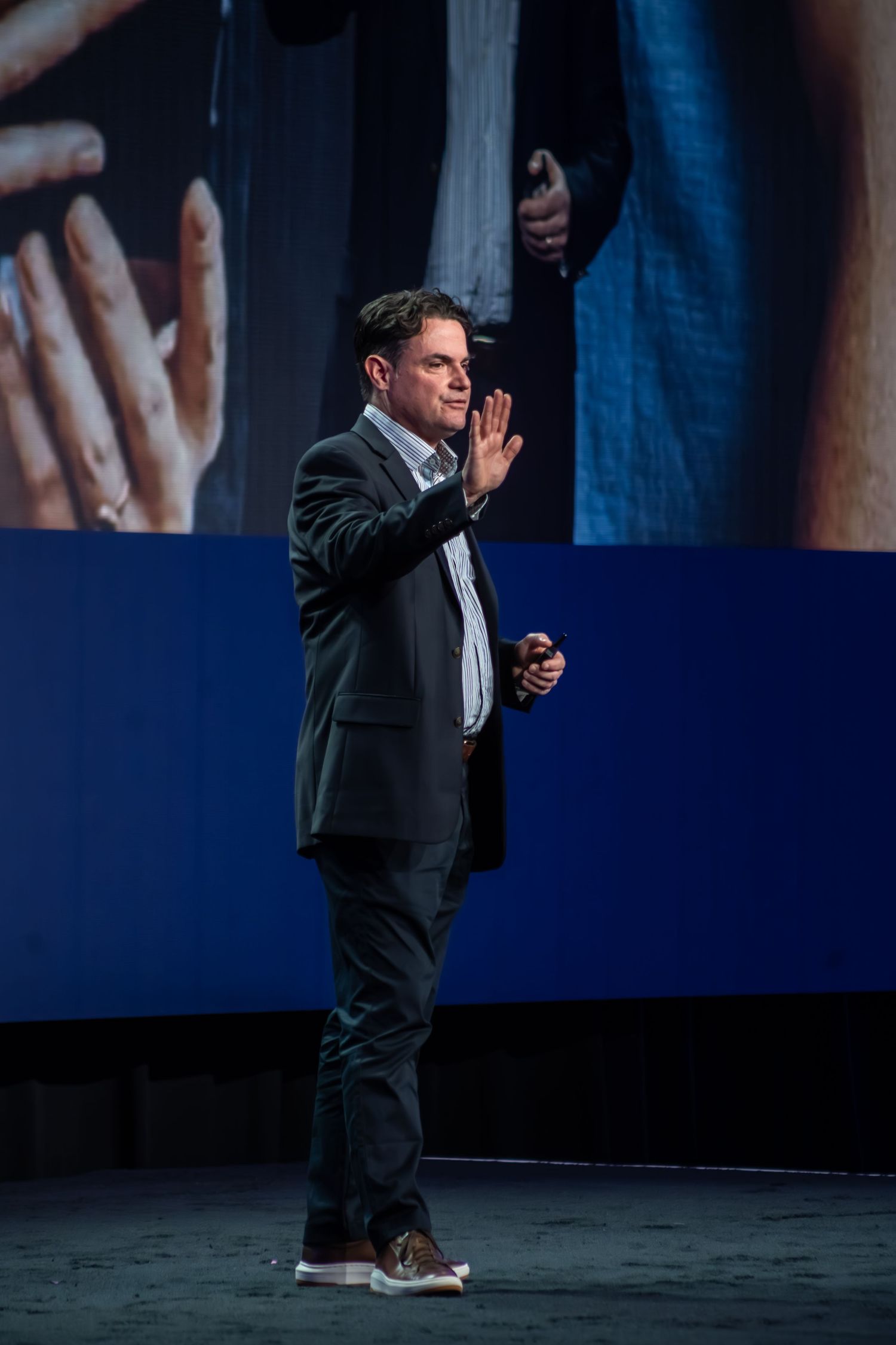Man in dark suit gesturing on stage during a conference presentation with blue screen backdrop.
