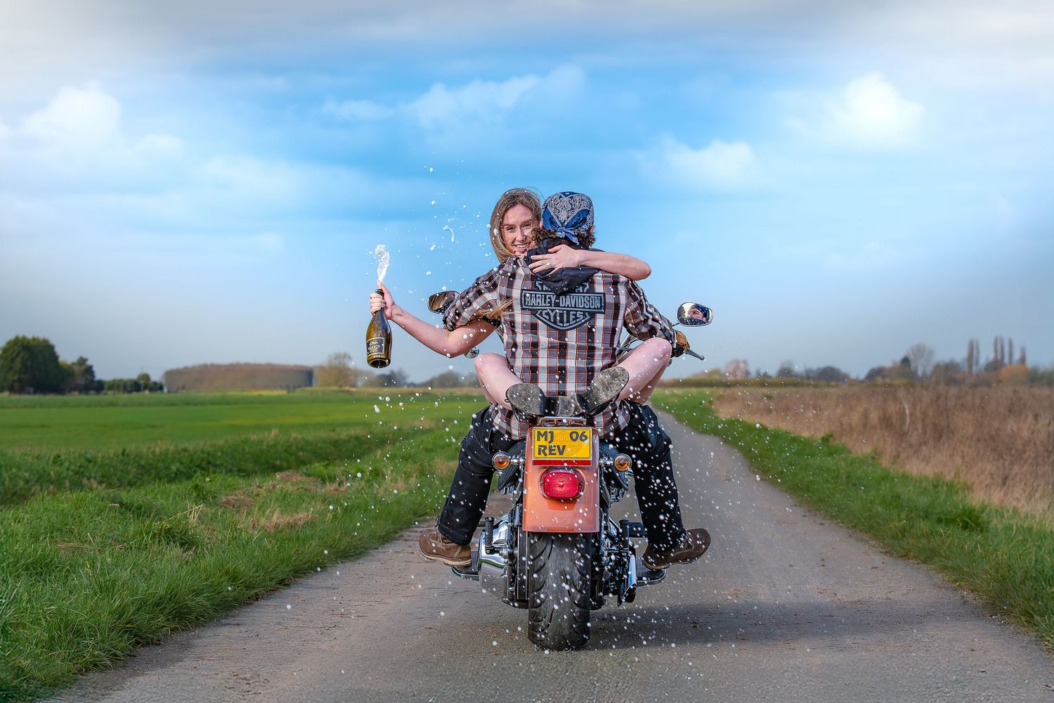 Couple riding a motorcycle on a rural road, woman holding champagne bottle with liquid splashing, green fields and blue sky.