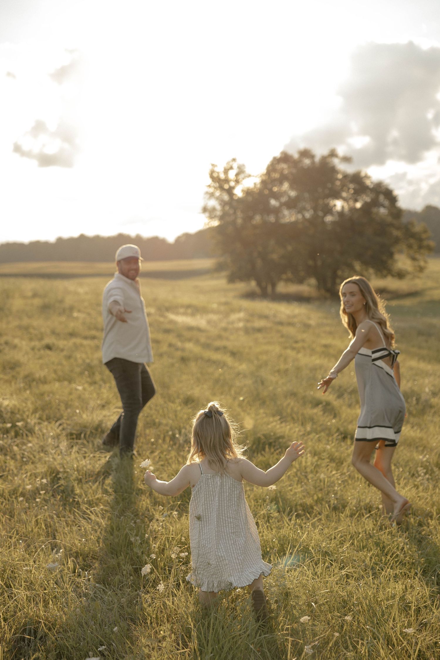 Family of three playing in a sunlit meadow at golden hour, with parents reaching out to their young daughter.