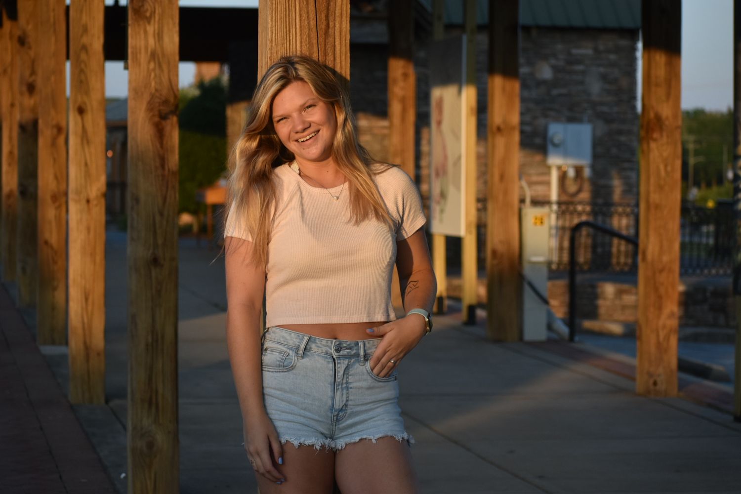 Young blonde woman smiling in white crop top and denim shorts, posing outdoors at golden hour.