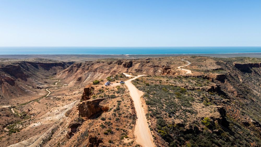 Aerial view of a winding dirt road through rugged canyon terrain leading to a vast blue ocean horizon.