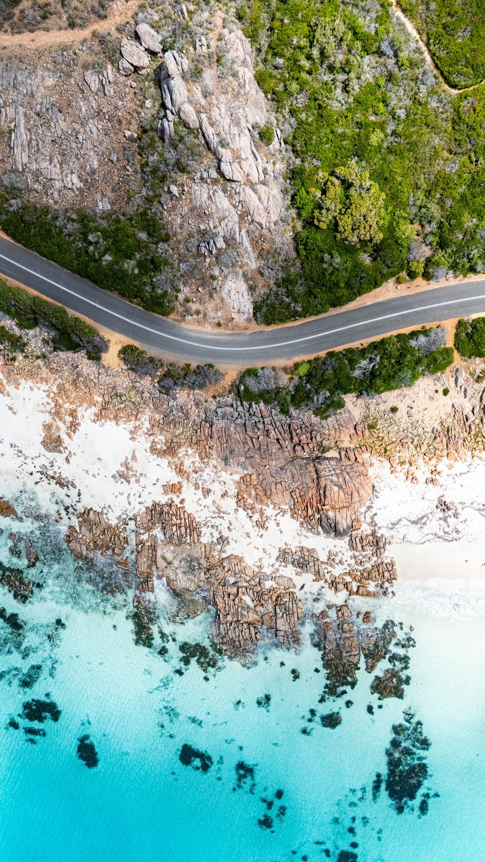 Aerial drone view of a winding coastal road between rocky cliffs and turquoise ocean waters with white sandy beach.
