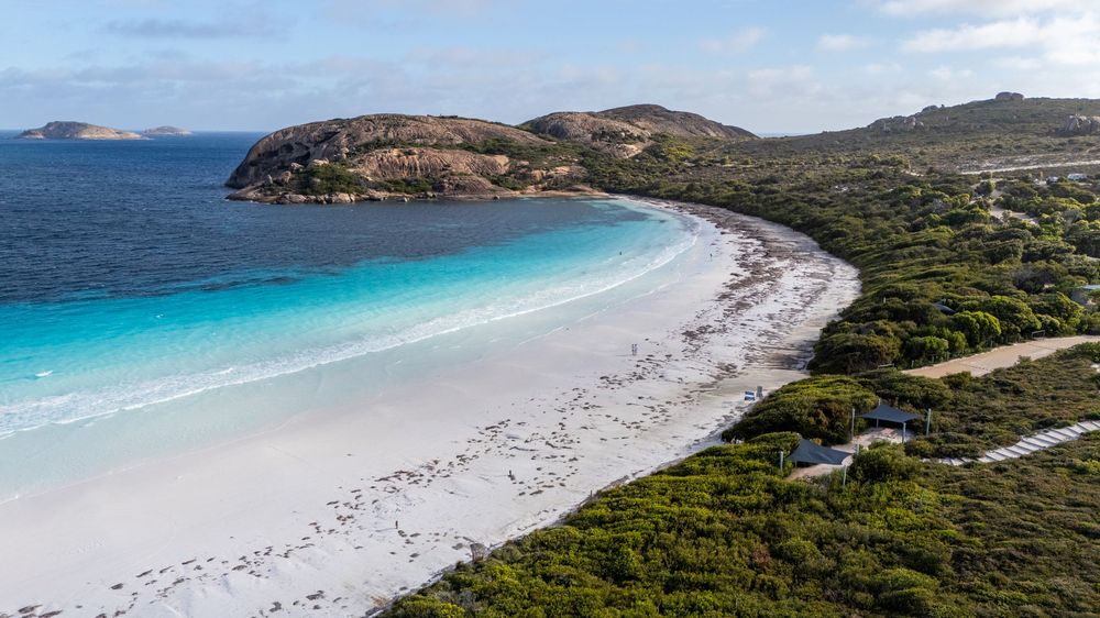 Aerial view of a pristine white sand beach with turquoise water, bordered by lush green bushland and rocky hills.