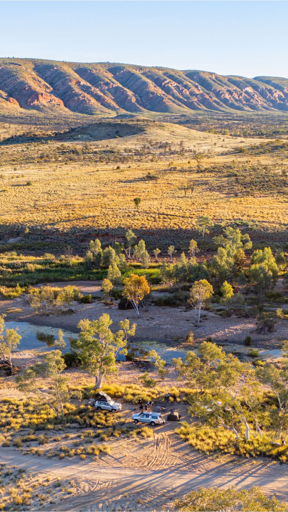Aerial view of Australian outback landscape with red mountains, arid plains, sparse trees, and two vehicles parked near a dry riverbed.