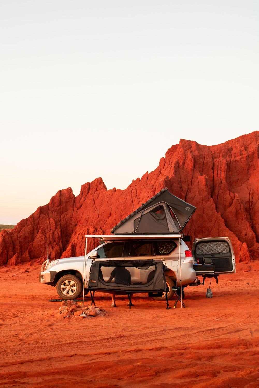 4WD vehicle with rooftop tent camped on red desert terrain against dramatic red rock cliffs at dusk.