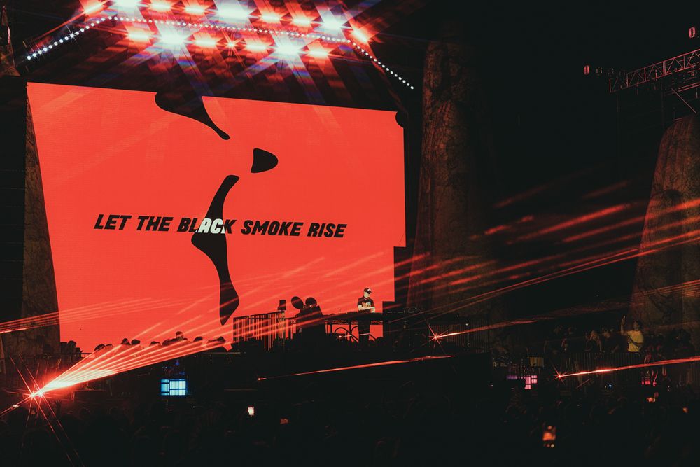 Concert stage with red screen displaying "Let The Black Smoke Rise" with laser lights at a live music event.