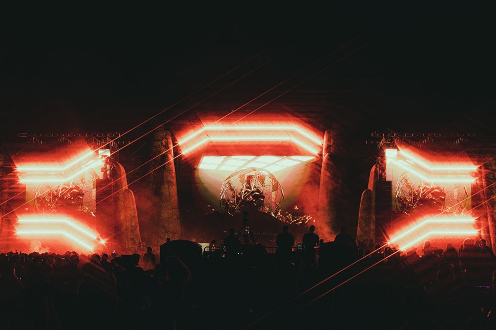 Massive outdoor music festival stage glowing with bright red neon lights and pyrotechnics against a dark night sky.