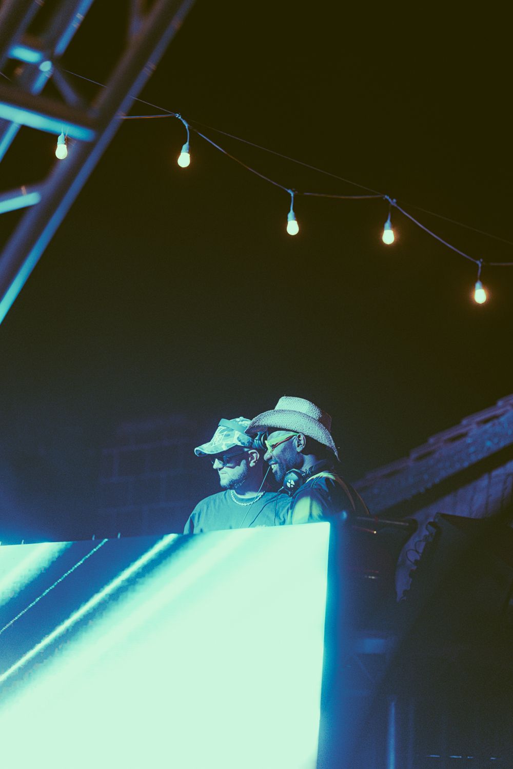 Two DJs wearing cowboy hats perform at an outdoor night event under string lights on a glowing blue-lit stage.
