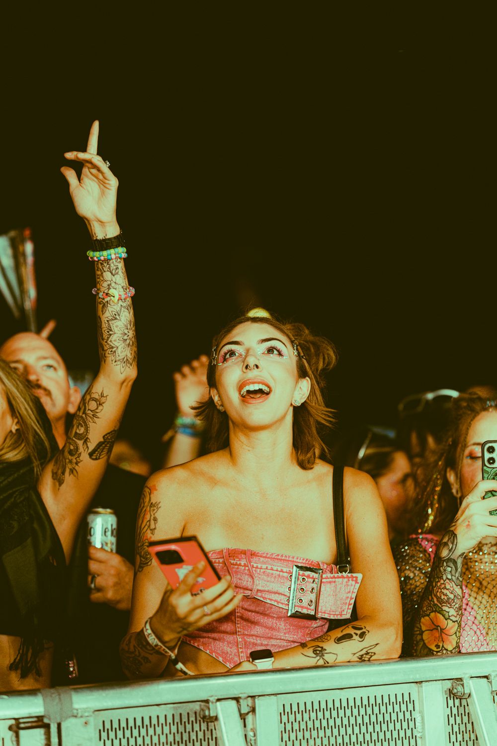 Tattooed woman in pink corset top holding phone and pointing up at outdoor music festival concert crowd.