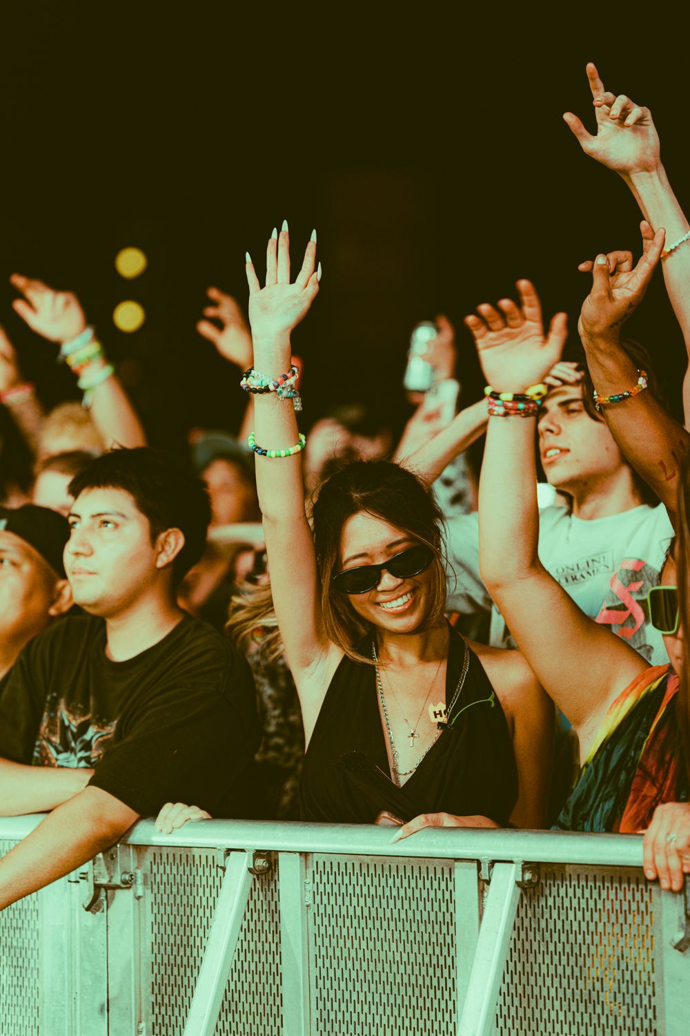 Excited crowd at a music festival with hands raised, wearing colorful bracelets, behind a metal barrier under green stage lighting.