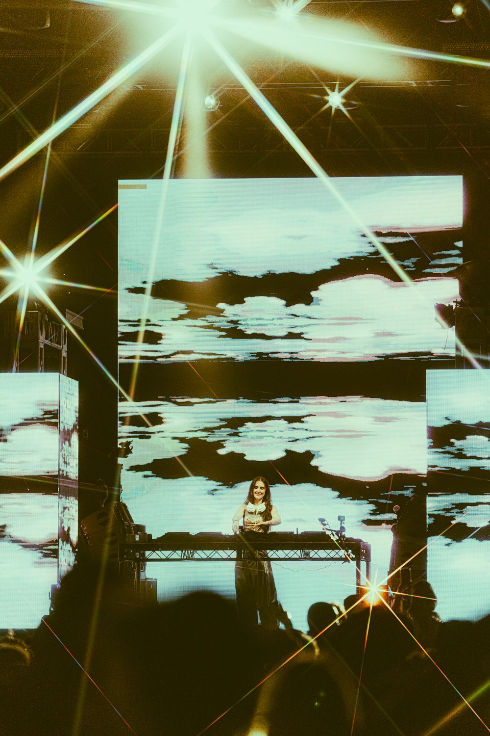 DJ performing on stage with large LED screens displaying clouds and bright star-shaped light beams at a concert.