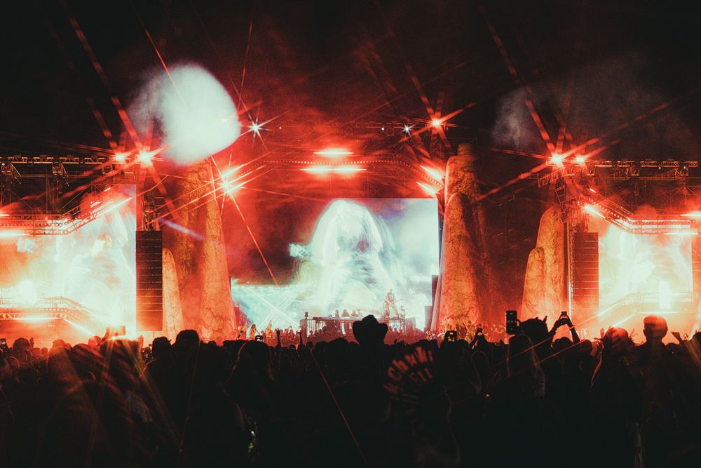 Crowd at an outdoor concert with dramatic red and orange stage lighting, laser beams, and a large LED screen.