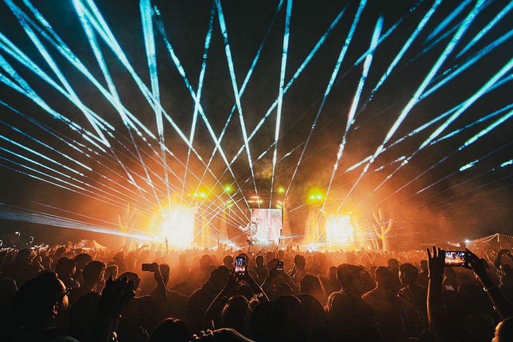 Crowd at an outdoor music festival with blue laser lights and glowing orange stage lights at night.