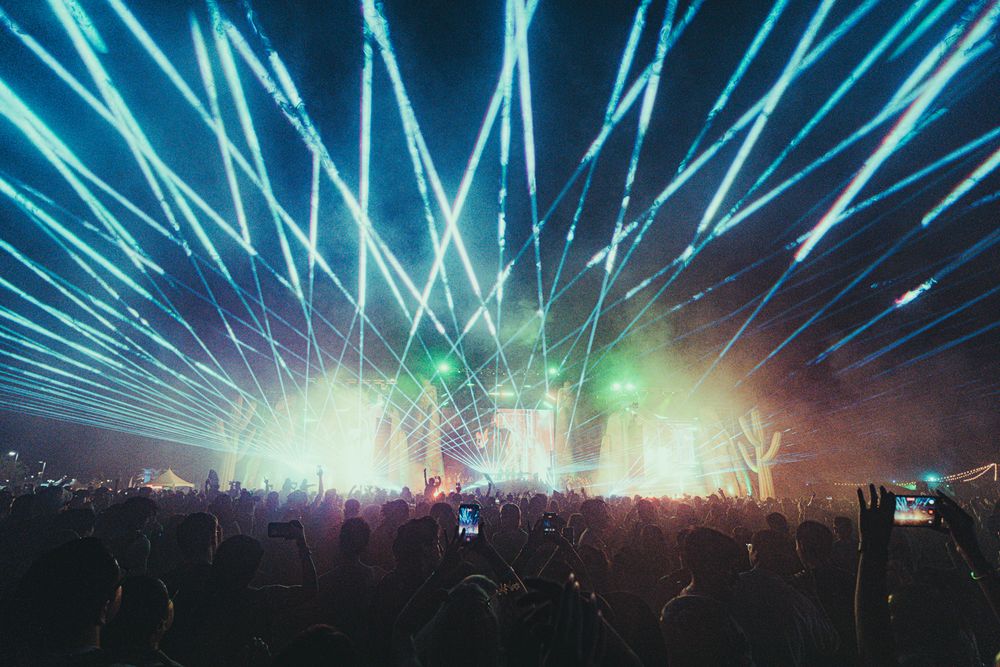Massive outdoor music festival crowd with spectacular blue laser light show and glowing stage at night.