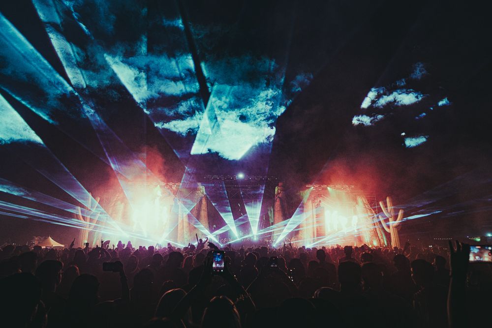 Dramatic outdoor concert stage with colorful laser lights and orange explosions illuminating a massive crowd at night.