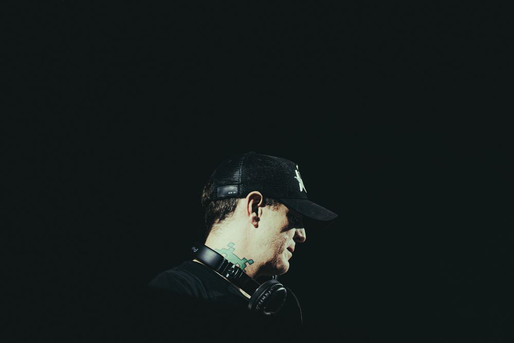 A tattooed DJ wearing a black cap and headphones around his neck poses in profile against a dark background.