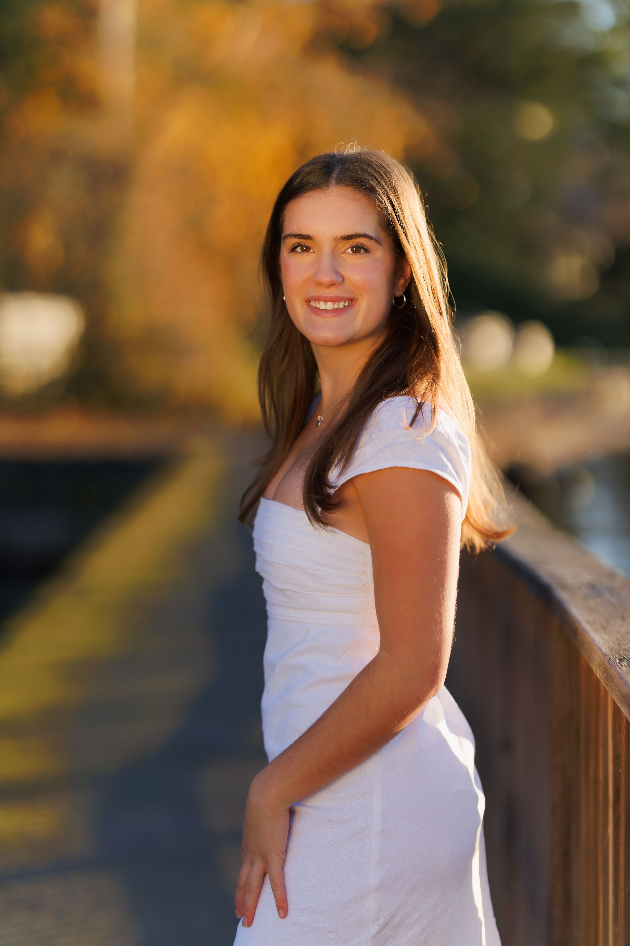 Young woman in white dress smiling on bridge with autumn foliage backdrop in golden hour light. Professional senior portrait.