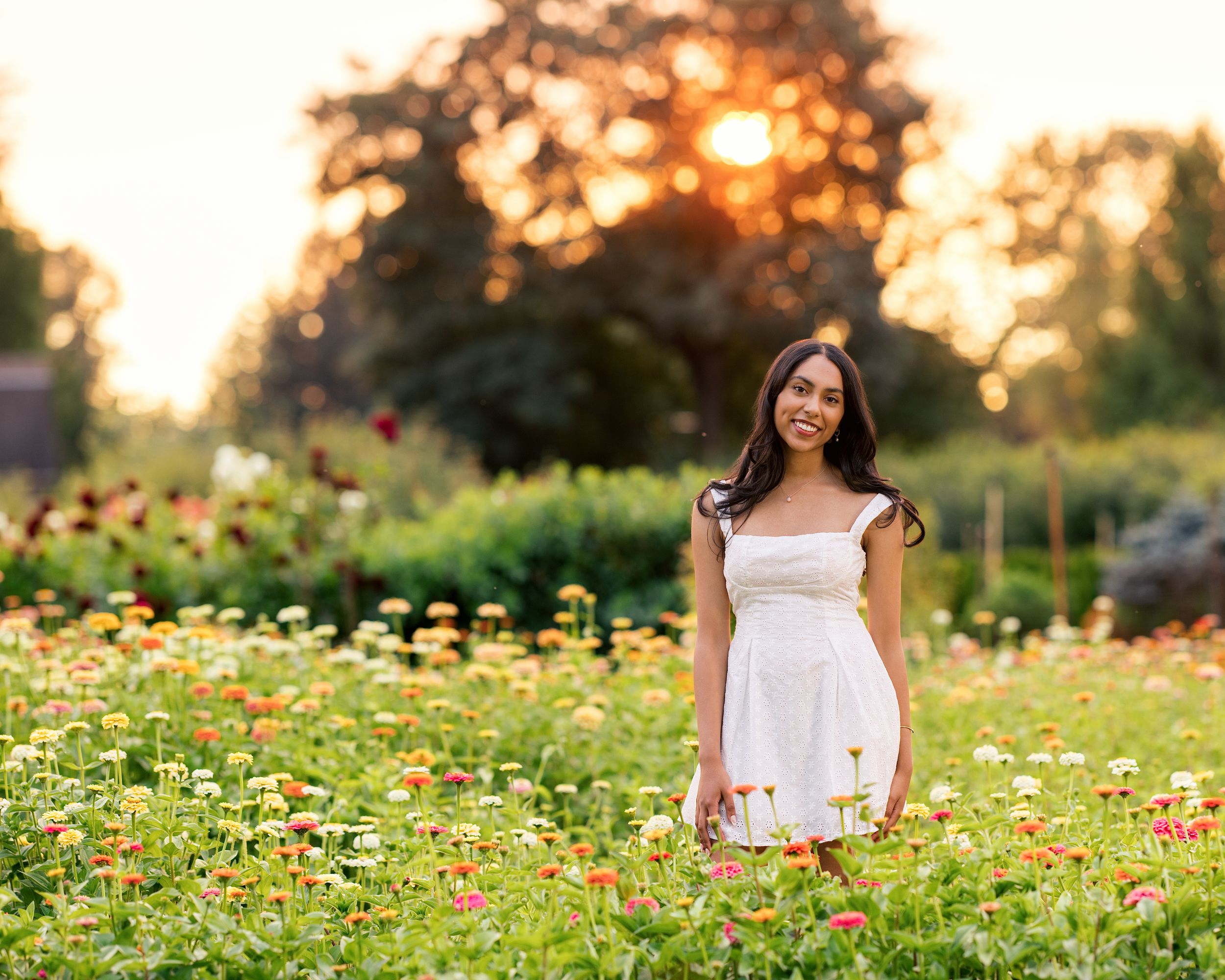 Young woman in white dress standing in colorful flower field at golden sunset hour.