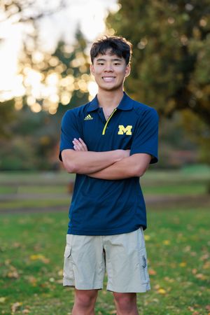 Young man in University of Michigan Adidas polo smiles with arms crossed outdoors in a park setting.