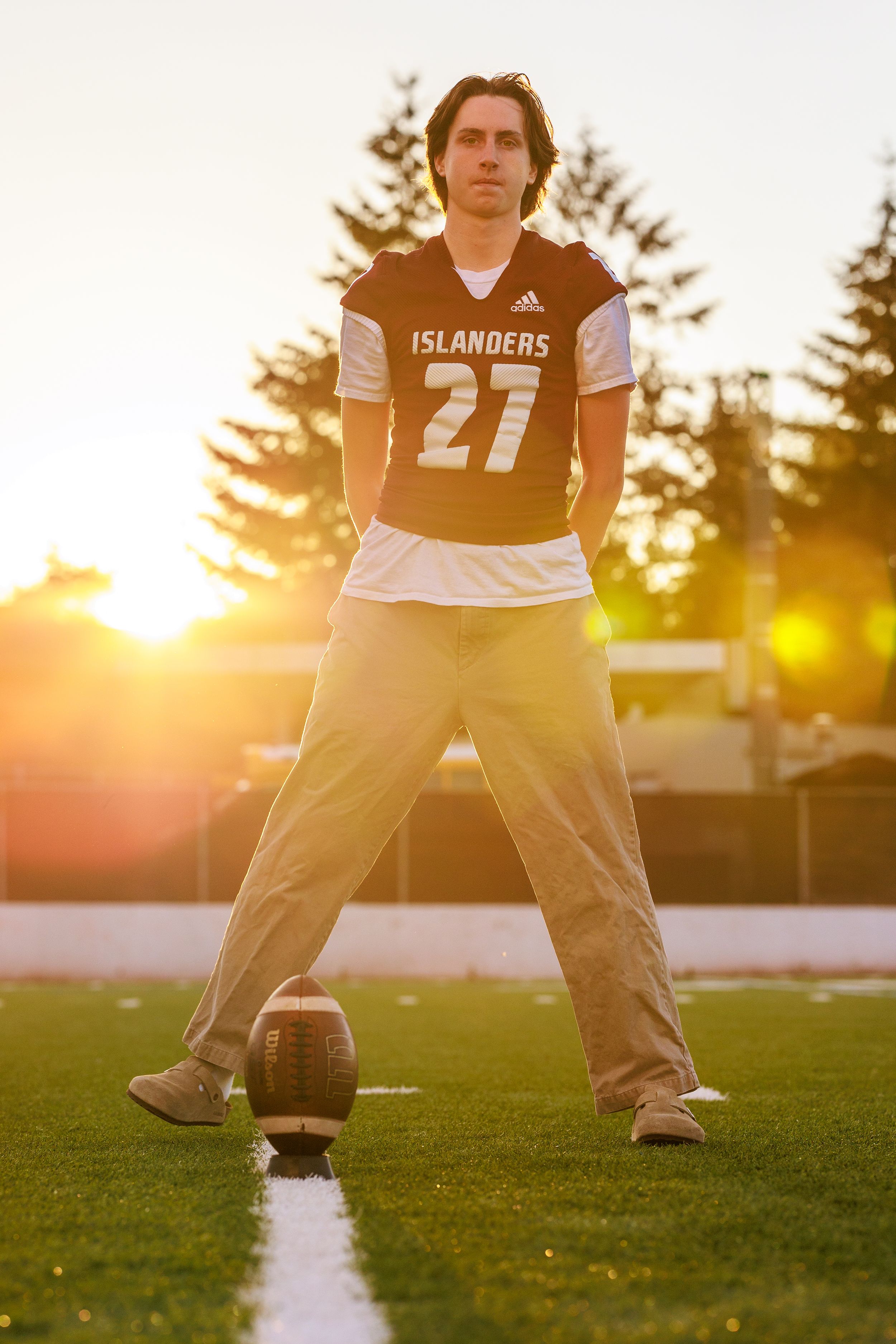 Young man in football jersey stands on turf field at sunset with football. Professional senior portrait.