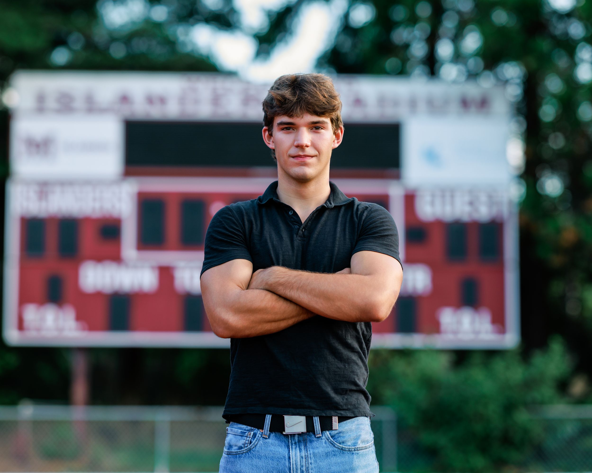 Young man in black polo shirt stands confidently with arms crossed in front of a football stadium scoreboard.