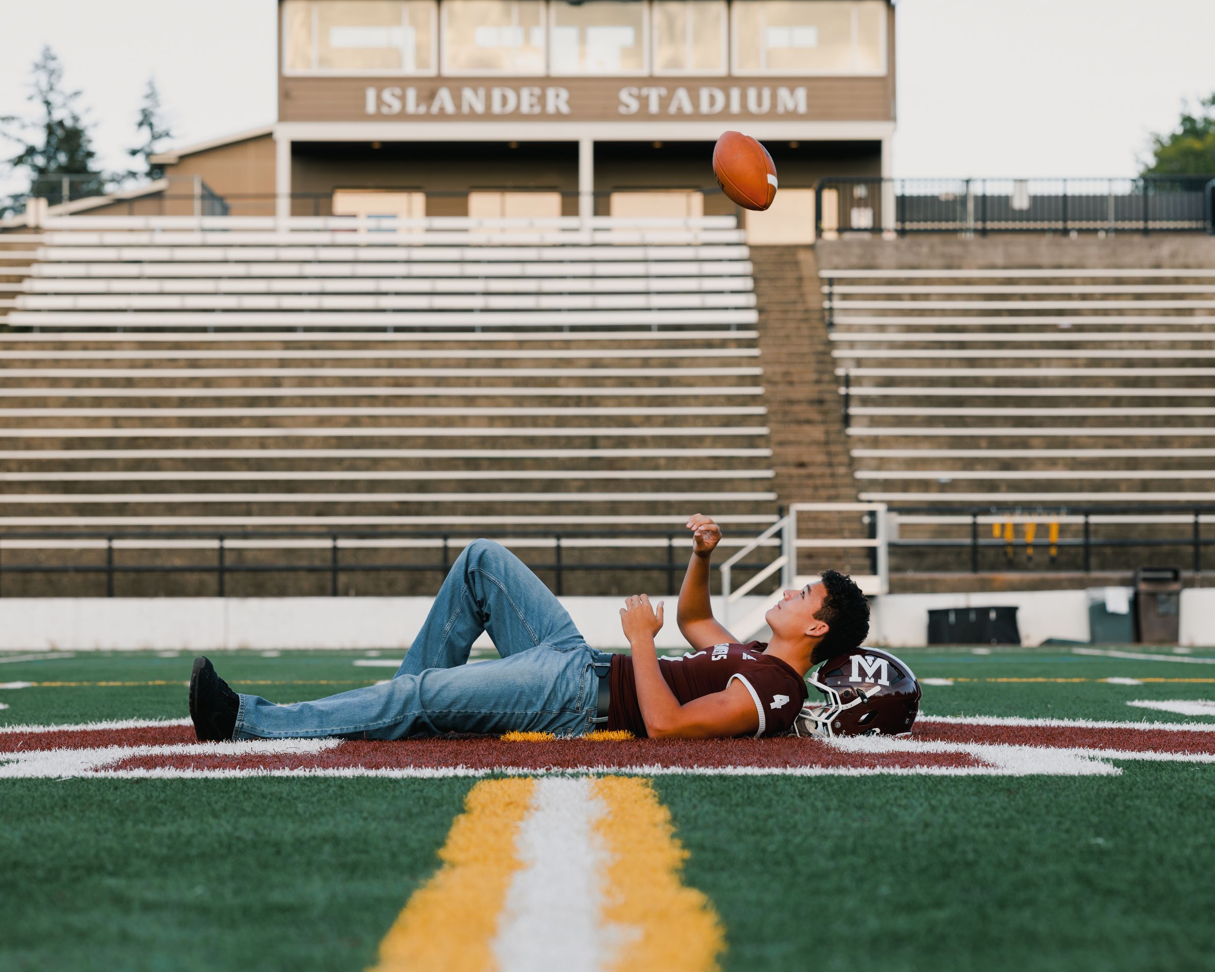 Teen lying on football field tossing a ball at Islander Stadium during a senior portrait session.