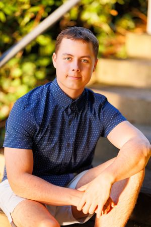 Young man in navy blue polka dot shirt sitting on outdoor steps with green foliage background. Professional senior portrait.