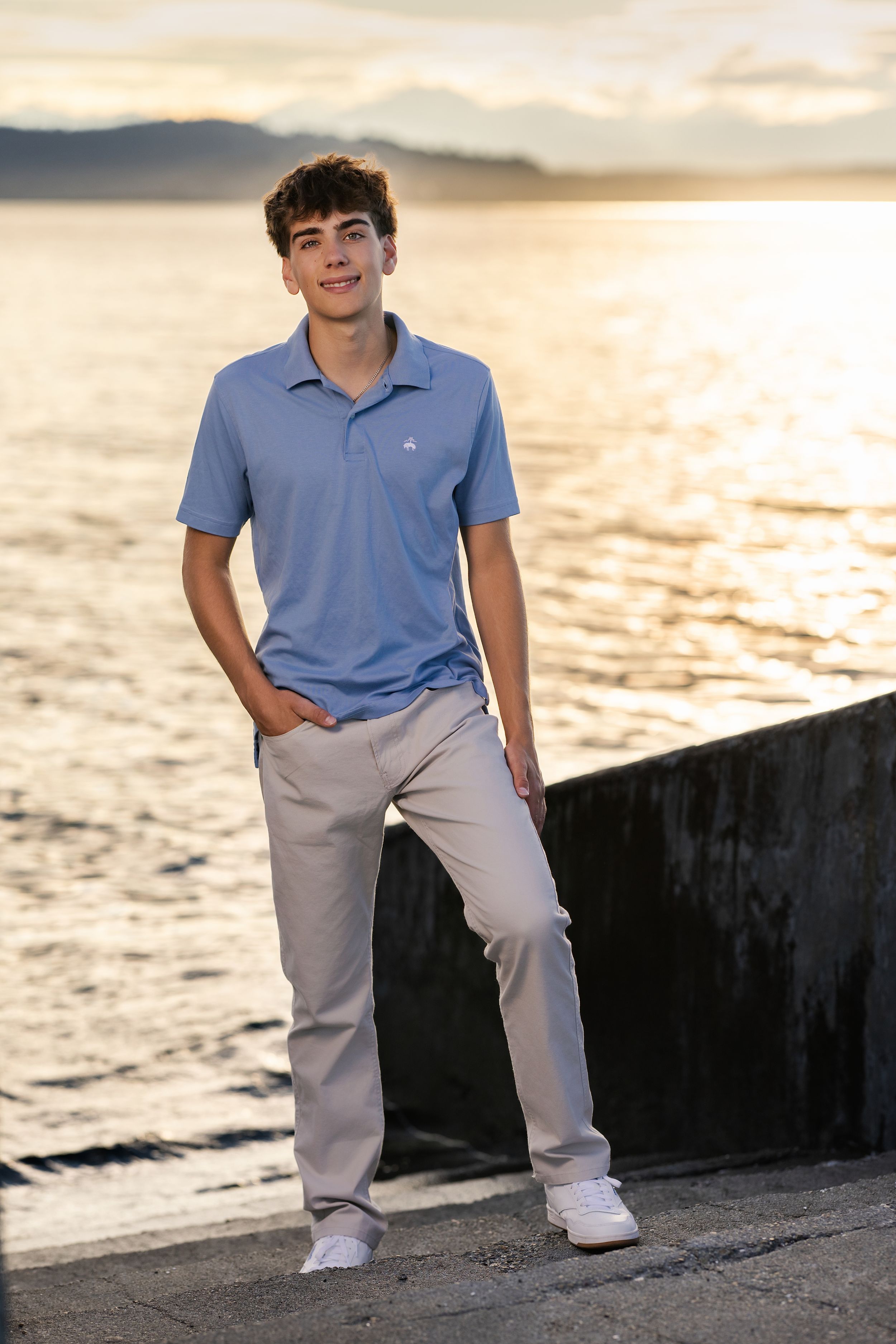 Teen boy in blue polo and khaki pants poses by waterfront wall at golden hour sunset. Professional senior portrait.