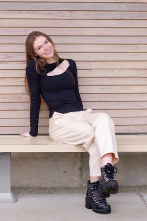 Young woman smiling on bench, wearing black top, cream pants, and black boots against wooden wall backdrop. Professional senior portrait.