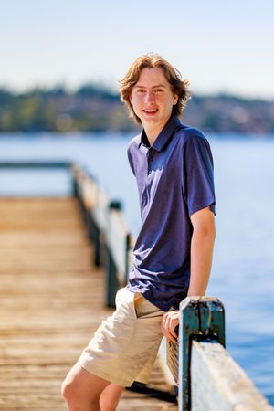 Smiling teenage boy in blue polo shirt and khaki shorts leaning on a wooden dock railing by a lake. Professional senior portrait.