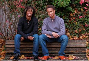 Two young men sitting together on a wooden bench outdoors, surrounded by colorful autumn foliage.