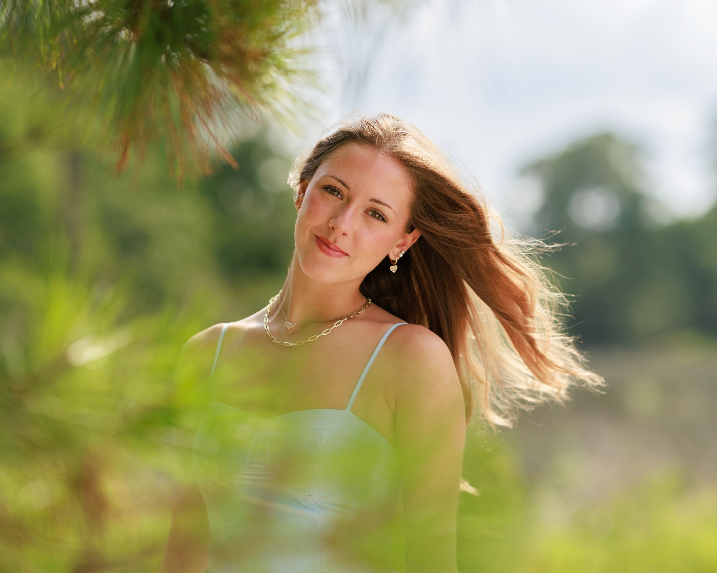 Young woman with flowing hair smiling outdoors among lush green trees in bright sunlight. Professional senior portrait.