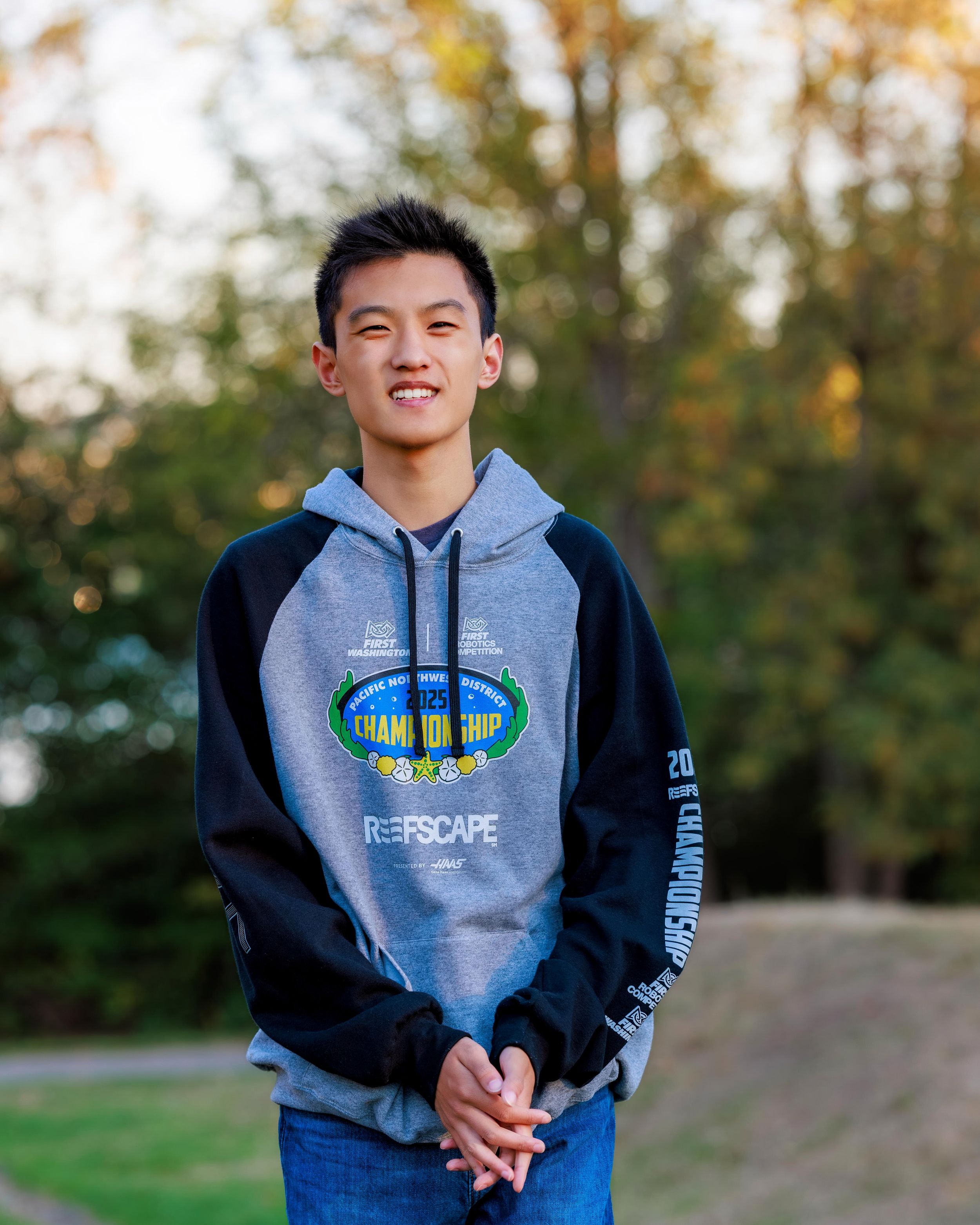 Smiling young boy wearing a gray and black Championship hoodie standing outdoors with trees in the background. Professional senior portrait.
