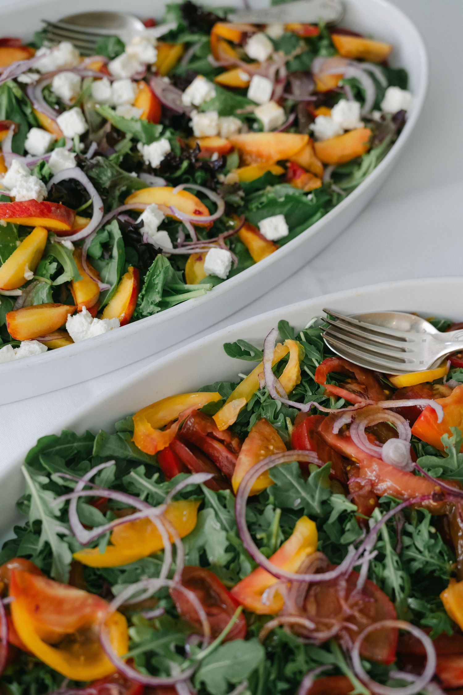 Two white oval platters of fresh arugula salad with peaches, red onion, bell peppers, and crumbled feta cheese.