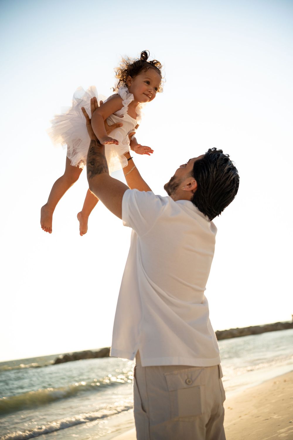 Father lifting happy toddler girl in white dress high in the air on a sunny beach.