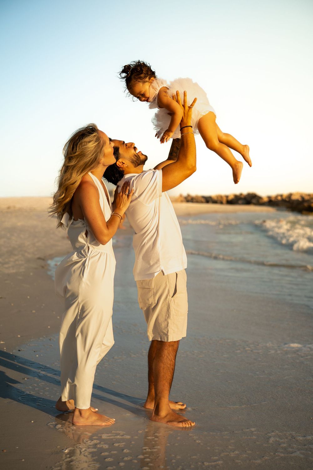 A joyful family in white outfits on a sunny beach, with a father lifting a baby while mother watches lovingly.