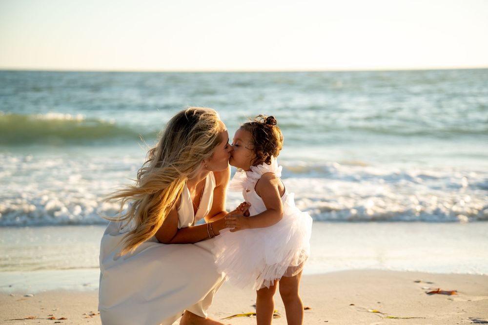 A mother holds her toddler daughter on a sunny beach, sharing a tender kiss near the ocean waves.