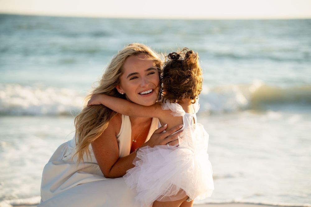 A smiling blonde woman hugs a young girl in a white dress on a sunny beach with ocean waves behind them.