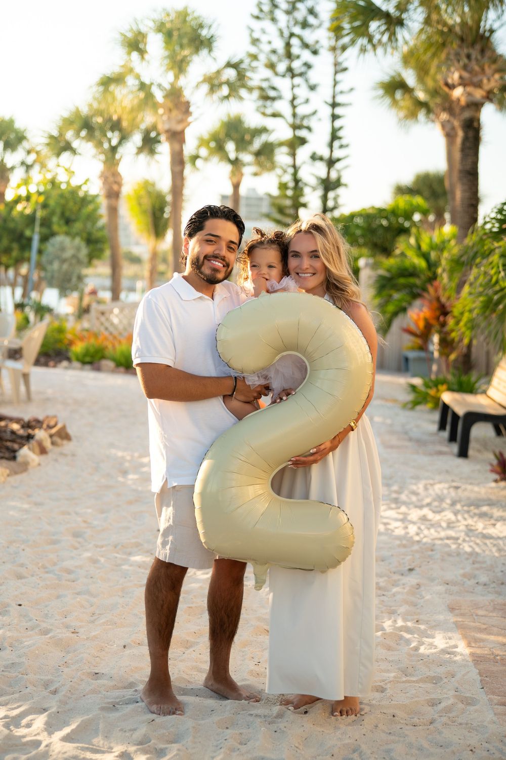 Family of three celebrating a 2nd birthday with a gold number balloon on a sandy beach path lined with palm trees.