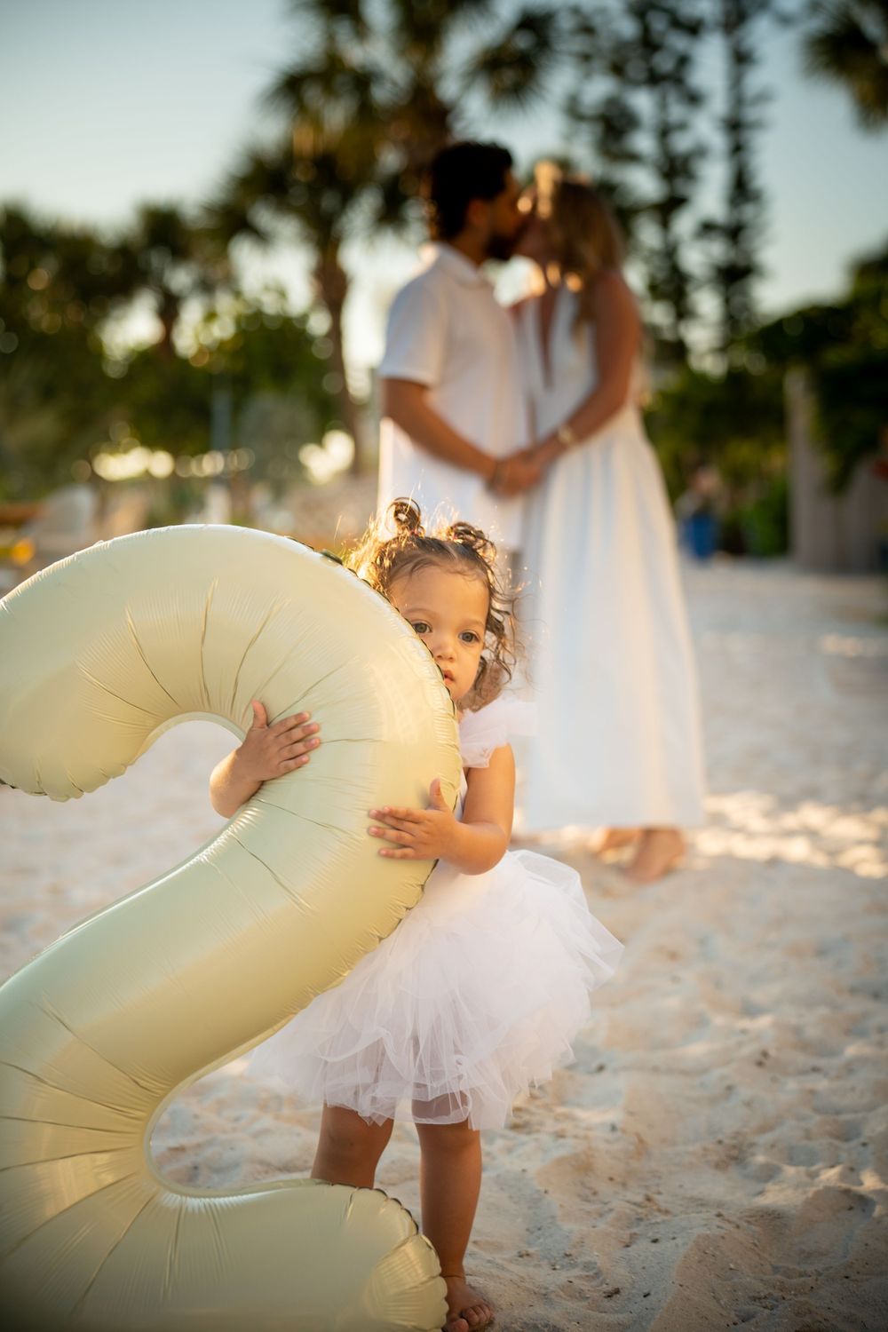 Little girl in white tutu hugs inflatable number 2 on beach while couple kisses in background at sunset.