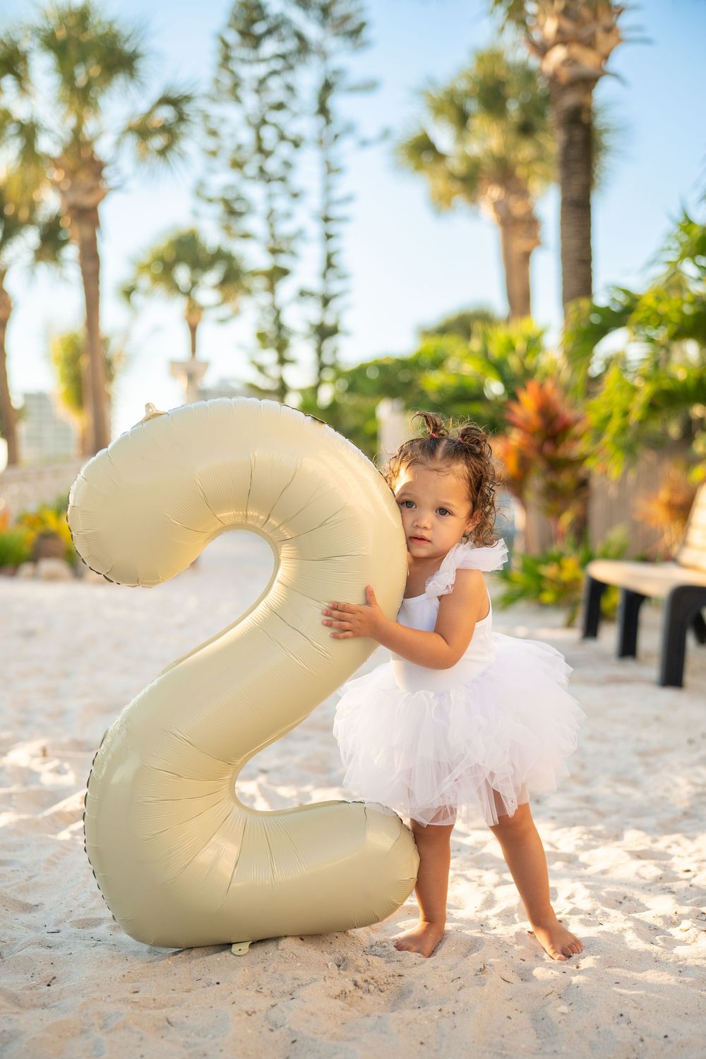Toddler girl in white tutu dress hugging a gold number 2 balloon on a sandy beach with palm trees.