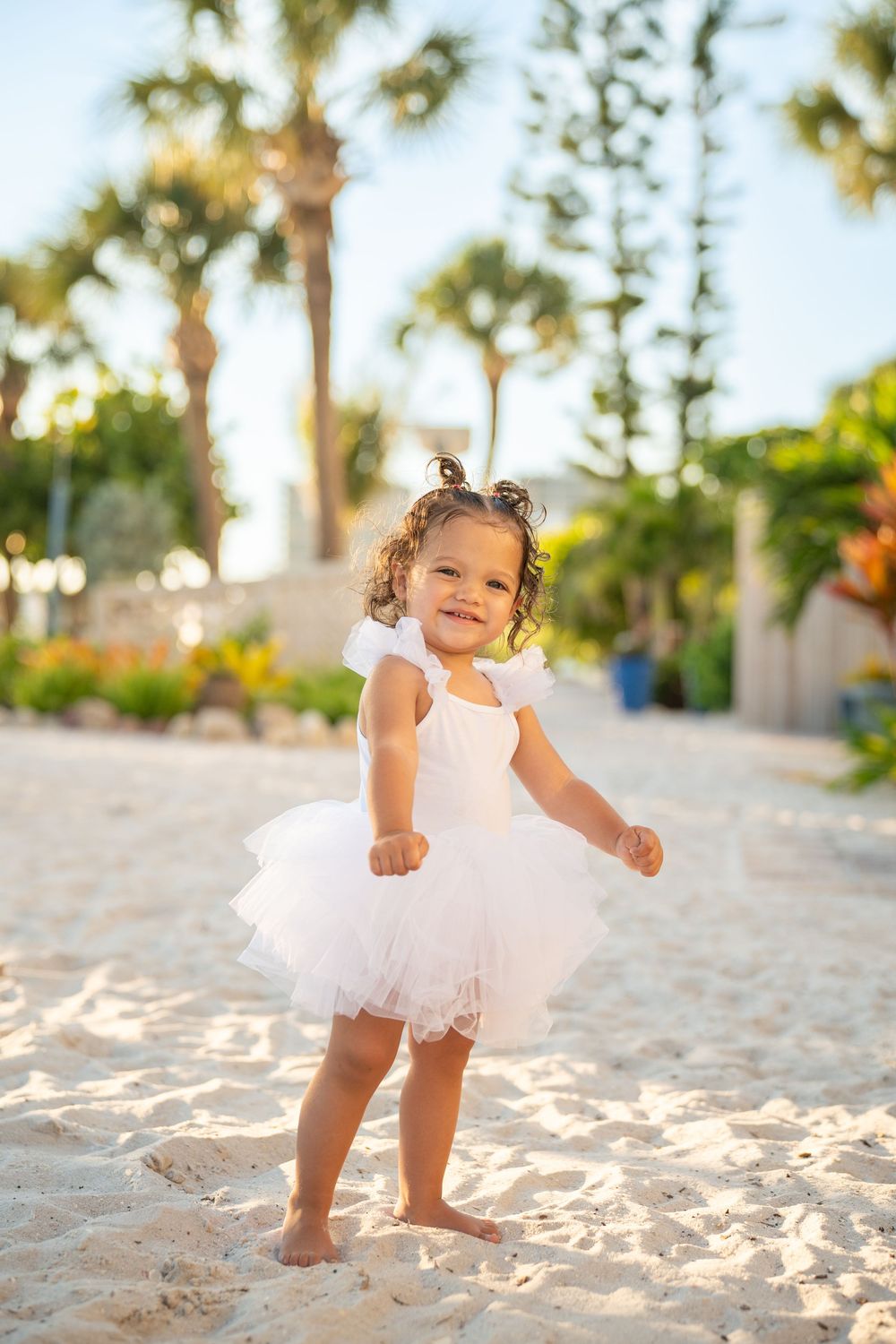 Smiling toddler girl in white tutu dress playing barefoot on sandy beach with palm trees in background.