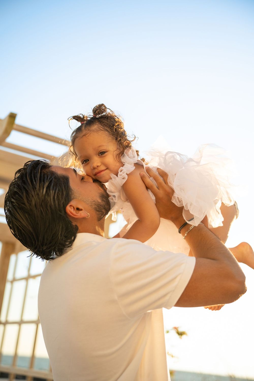 Father kissing his toddler daughter in a white dress, lifted in the air at sunset.