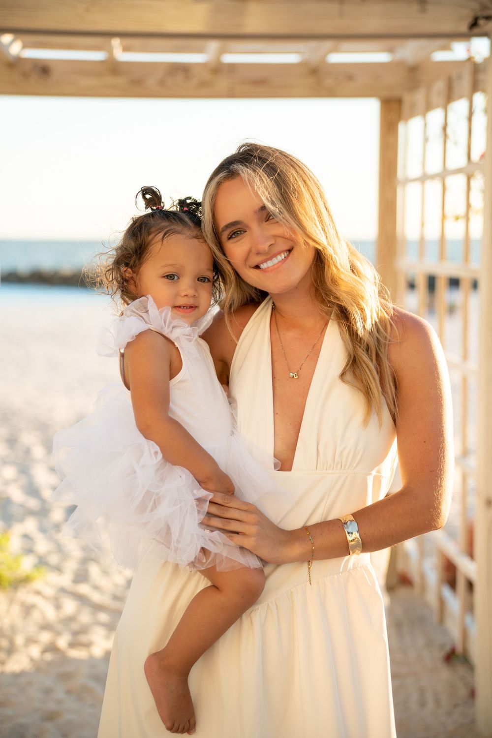 Smiling blonde mother in white dress holding her toddler daughter in a white tutu on a sunny beach.