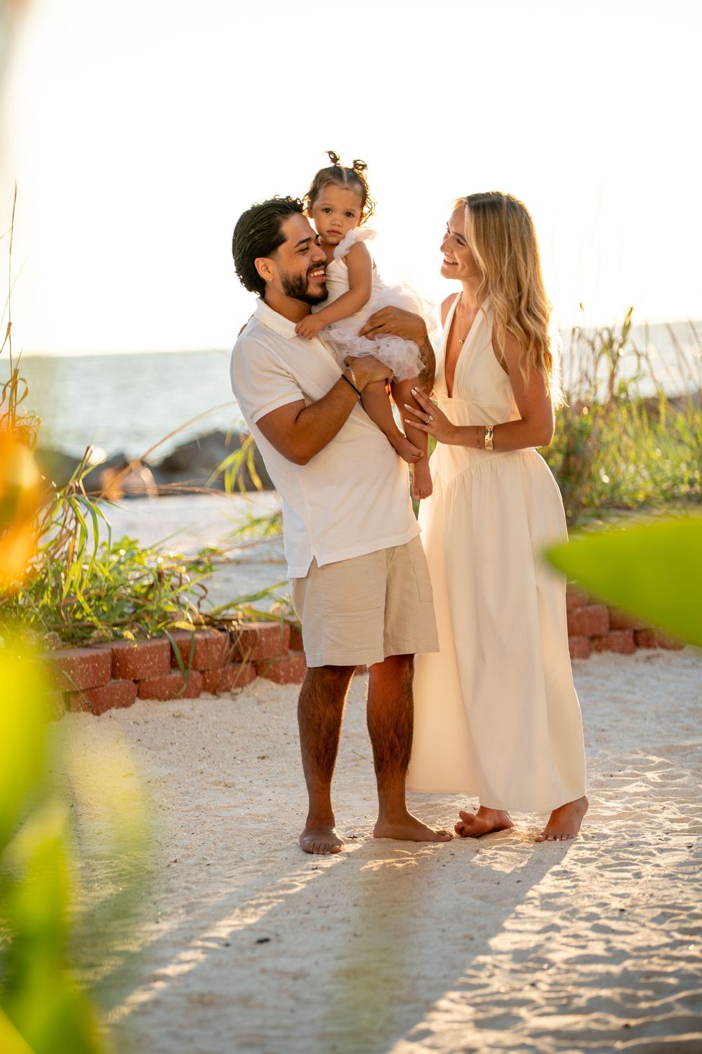 Happy family of three with parents in white outfits holding their toddler daughter on a sandy beach at sunset.