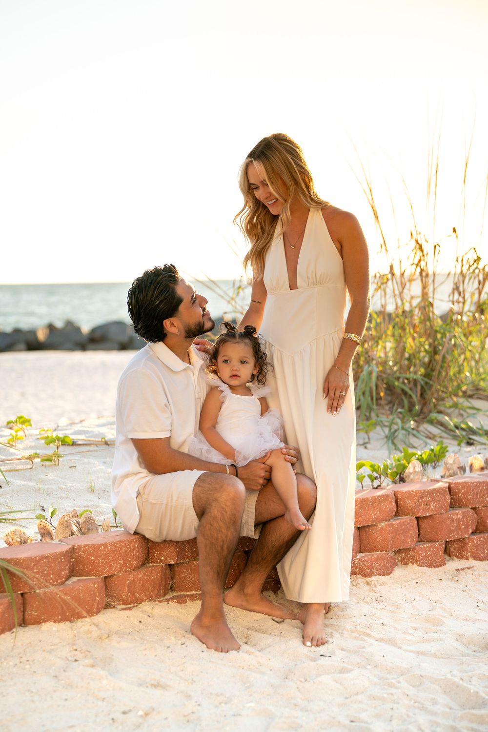 Family of three poses on brick wall at sandy beach, parents in white outfits smiling at toddler girl at sunset.