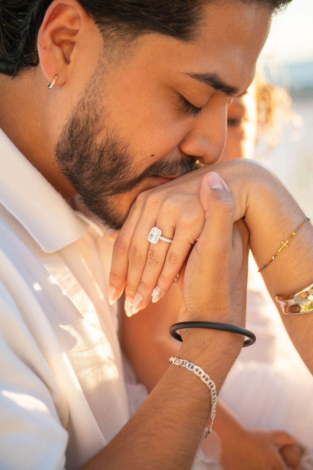 Man kissing woman's hand adorned with engagement ring, bracelets, and cross bracelet in romantic golden light.