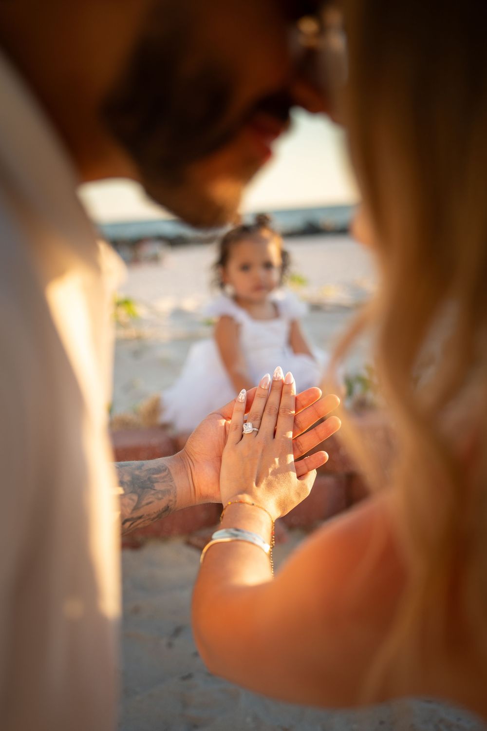 Couple holding hands showing engagement ring on beach at sunset with young girl in white dress in background.