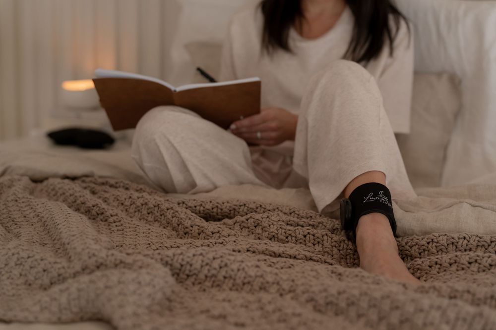 Woman in white pajamas sitting on a cozy knit blanket on bed, writing in a journal by warm candlelight.