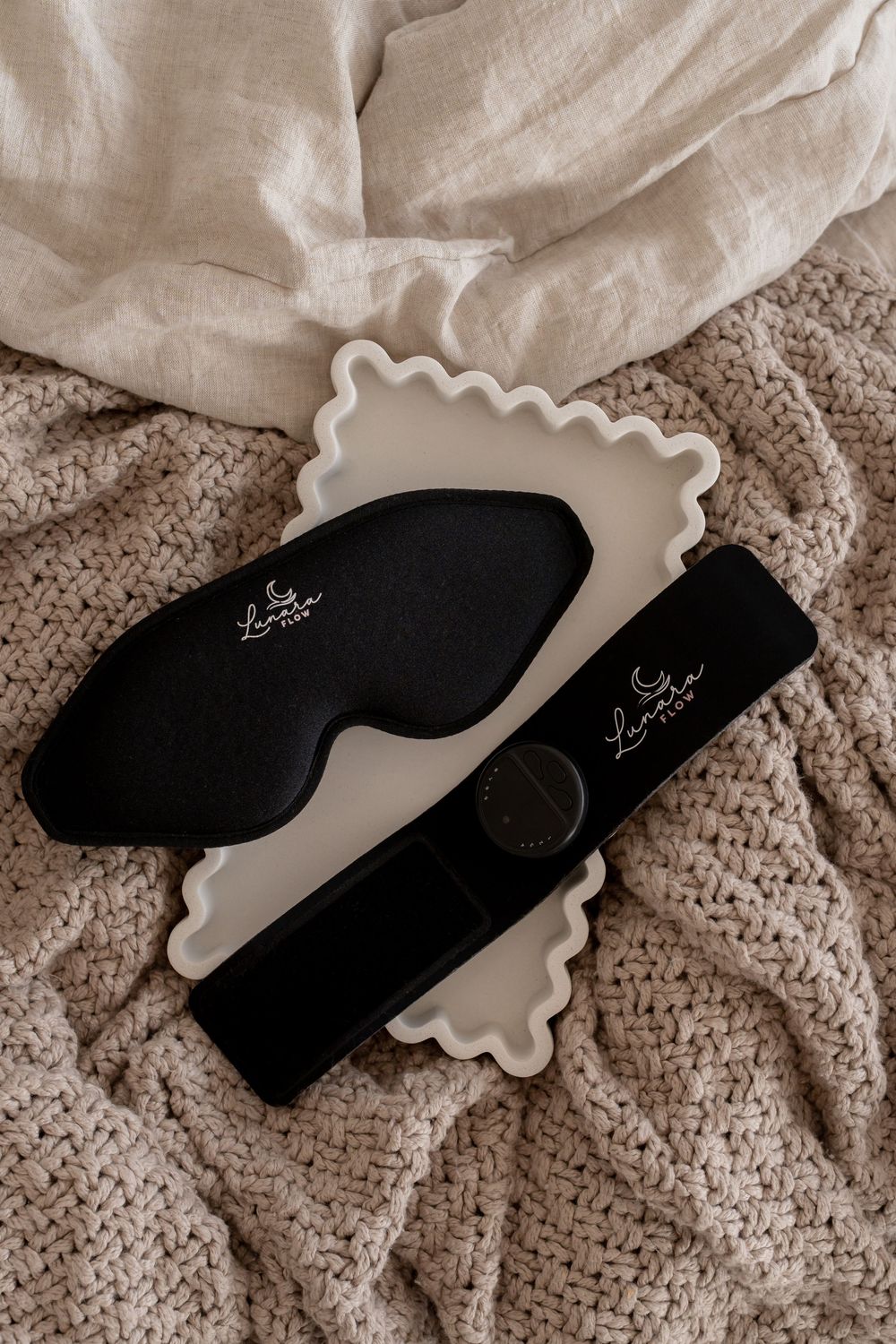 Black silk sleep mask and travel case on white scalloped tray atop cozy knit blanket, luxury bedtime accessories.